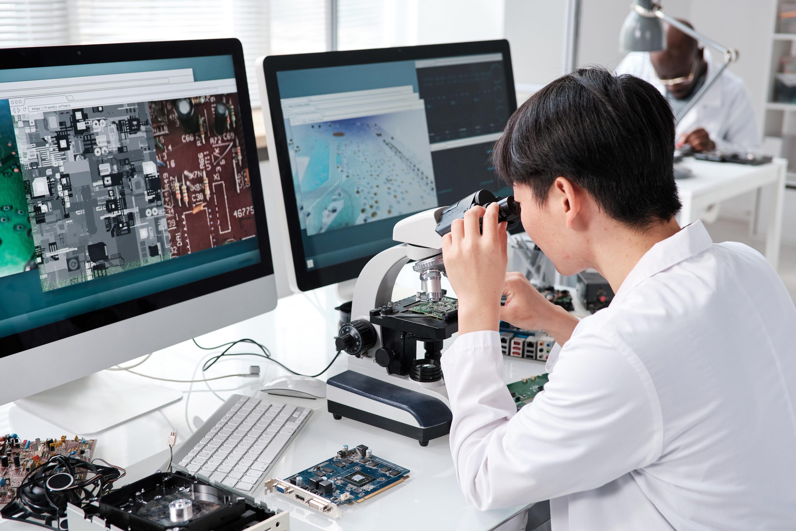 Engineer analyzing circuit boards through a microscope with inspection visuals displayed on monitors, representing the use of a deep learning vision system for quality control in electronics manufacturing.